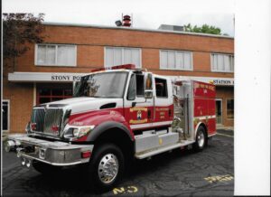 A red and white fire truck parked outside a brick fire station building labeled "STONY POINT STATION 1" on a sunny day. The fire truck has emergency lights and logos on its side.