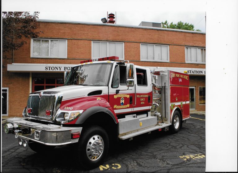 A red and white fire truck parked outside a brick fire station building labeled "STONY POINT STATION 1" on a sunny day. The fire truck has emergency lights and logos on its side.