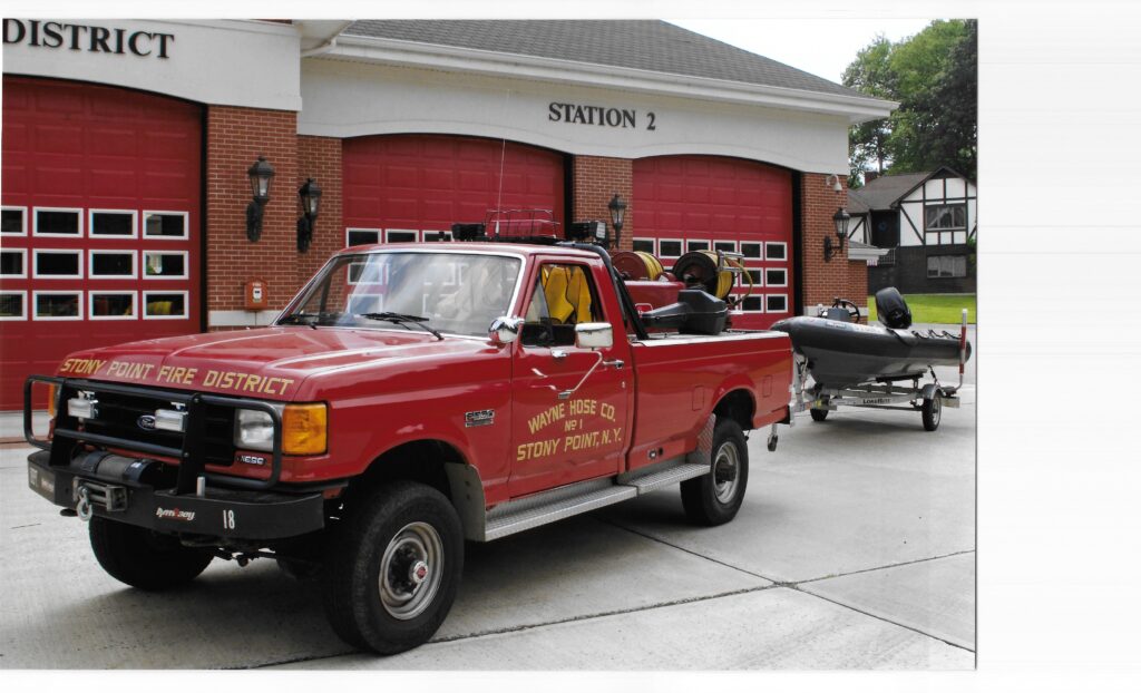 A red Stony Point Fire District truck is parked outside a fire station with red doors, towing a small rescue boat.