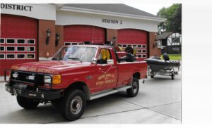 A red Stony Point Fire District truck is parked outside a fire station with red doors, towing a small rescue boat.
