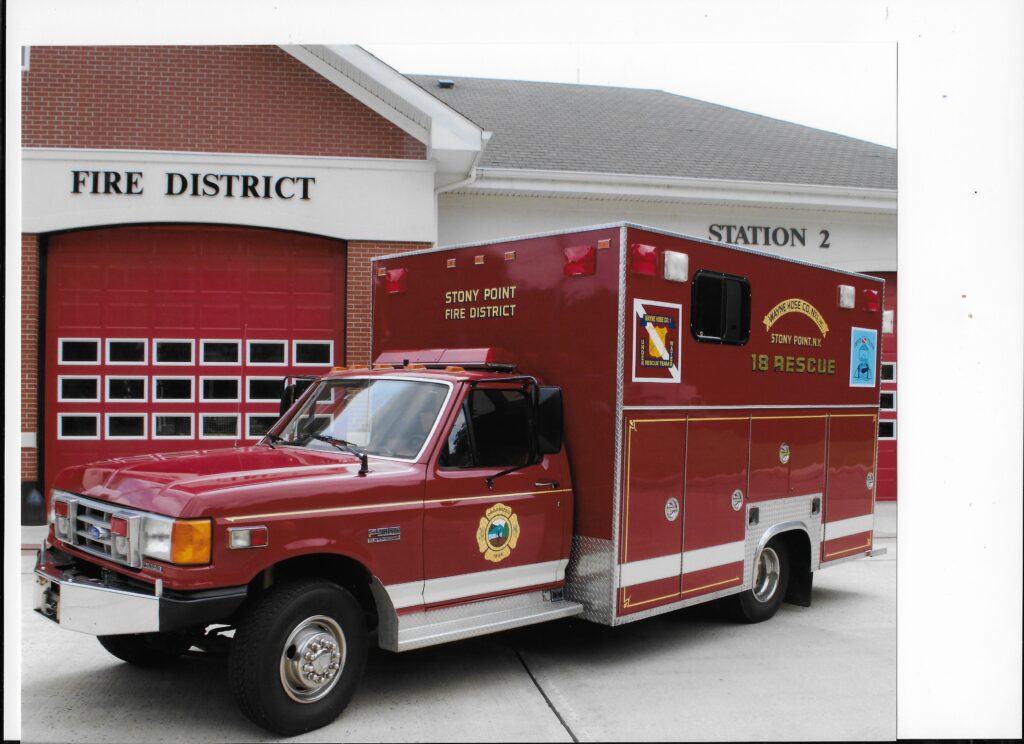 A red rescue vehicle labeled "Stony Point Fire District" is parked in front of a fire station with red garage doors and a sign reading "Fire District Station 2.