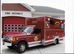 A red rescue vehicle labeled "Stony Point Fire District" is parked in front of a fire station with red garage doors and a sign reading "Fire District Station 2.