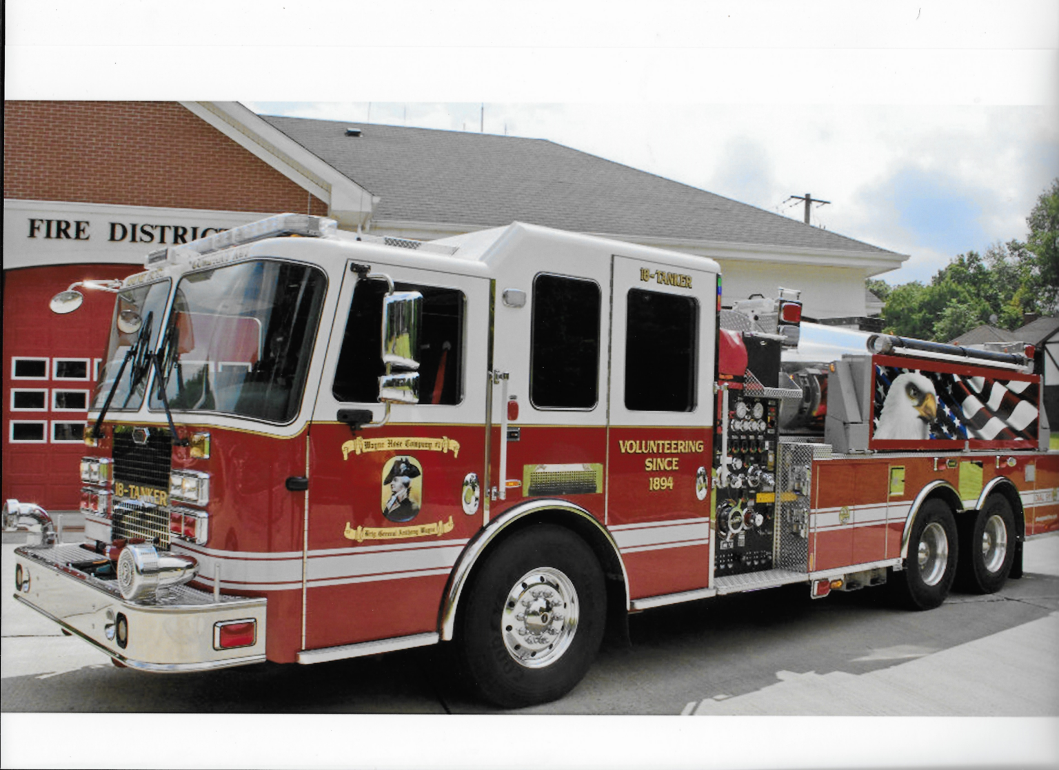 A red and white fire truck is parked outside a fire station with red bay doors. The truck has “VOLUNTEERING SINCE 1894” written on the side and is equipped with various hoses and equipment.