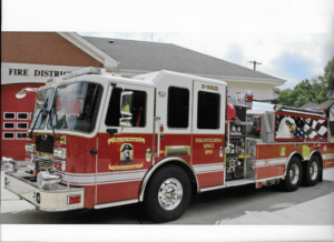 A red and white fire truck is parked outside a fire station with red bay doors. The truck has “VOLUNTEERING SINCE 1894” written on the side and is equipped with various hoses and equipment.