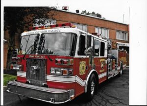 A red fire truck labeled "Stony Point N.Y." and "Pride of the Point" is parked outside a brick fire station, with trees and another vehicle visible in the background.