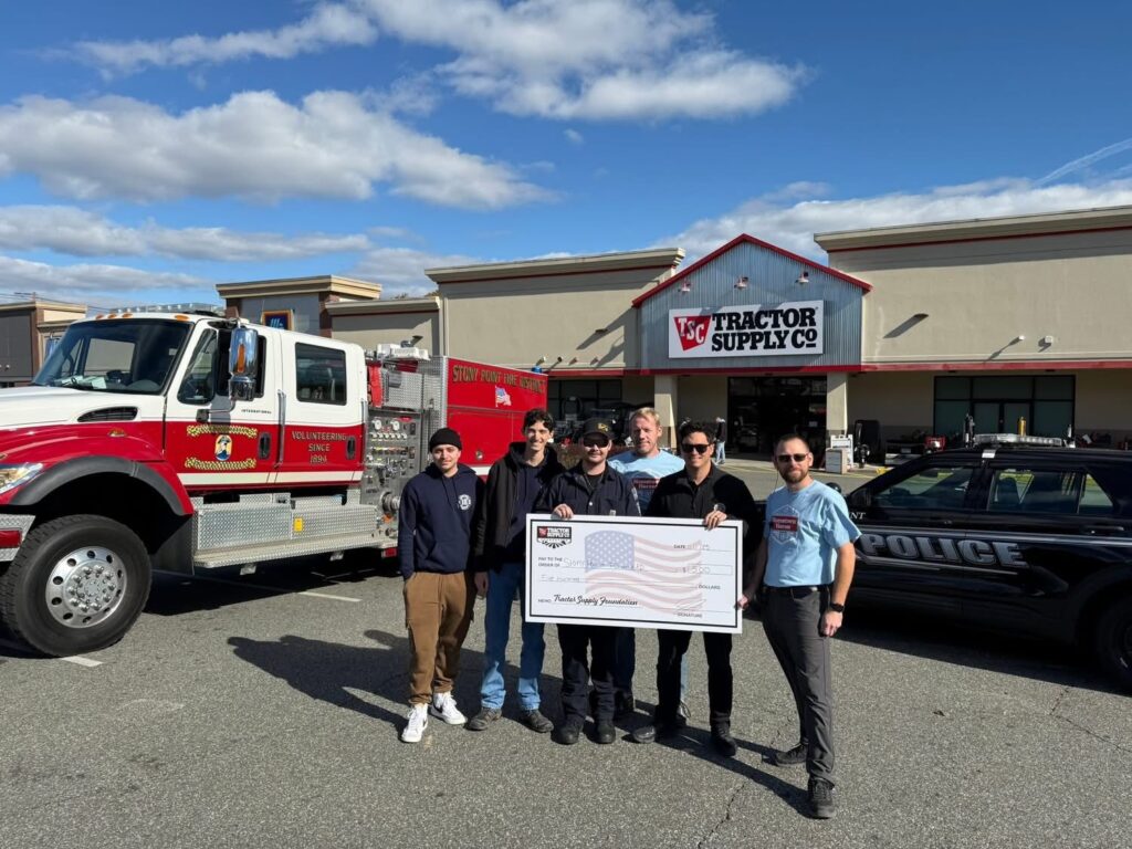 A group of people stand in front of a fire truck and police car outside a Tractor Supply Co. store, holding a large ceremonial check with an American flag design. The sky is blue with clouds.