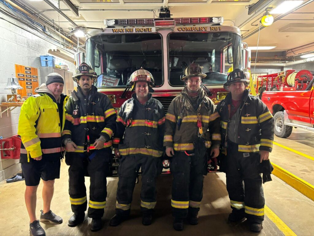Five people stand in front of a firetruck in a fire station. Four are dressed in firefighter gear and helmets, while one, on the left, wears a yellow jacket, black shorts, and a beanie. They are posing and smiling for the photo.