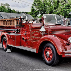 A vintage red fire truck with "Wayne Hose Co. No 1 Stony Point, N.Y." on the side is parked on a street, featuring shiny chrome details and wooden ladders, with houses and trees in the background.