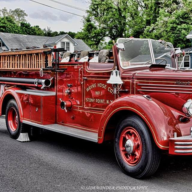 A vintage red fire truck with "Wayne Hose Co. No 1 Stony Point, N.Y." on the side is parked on a street, featuring shiny chrome details and wooden ladders, with houses and trees in the background.