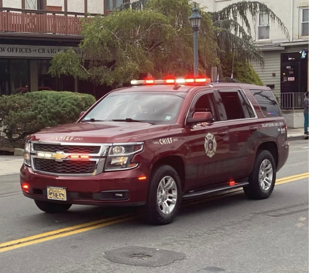 A maroon Chevrolet SUV with flashing red lights, marked "CHIEF" and "Fire Department," is parked on a street in front of buildings and greenery.