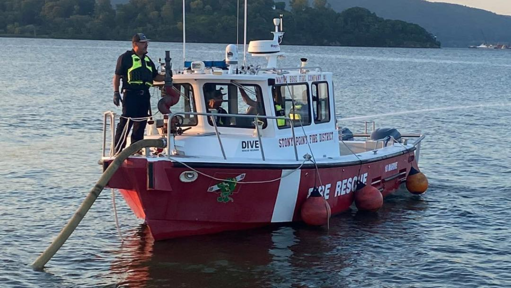 A red and white fire rescue boat labeled "DIVE" and "FIRE RESCUE" floats on calm water. Two crew members in safety gear stand on deck, and a large hose extends from the side of the boat into the water.