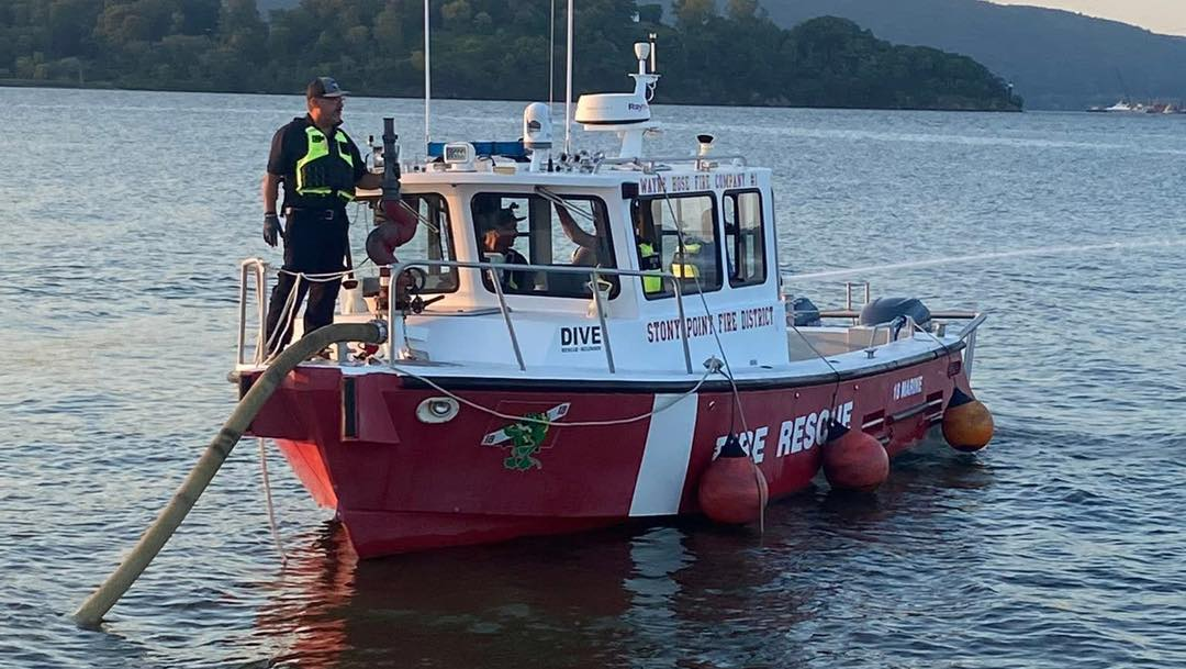 A red and white fire rescue boat labeled "DIVE" and "FIRE RESCUE" floats on calm water. Two crew members in safety gear stand on deck, and a large hose extends from the side of the boat into the water.