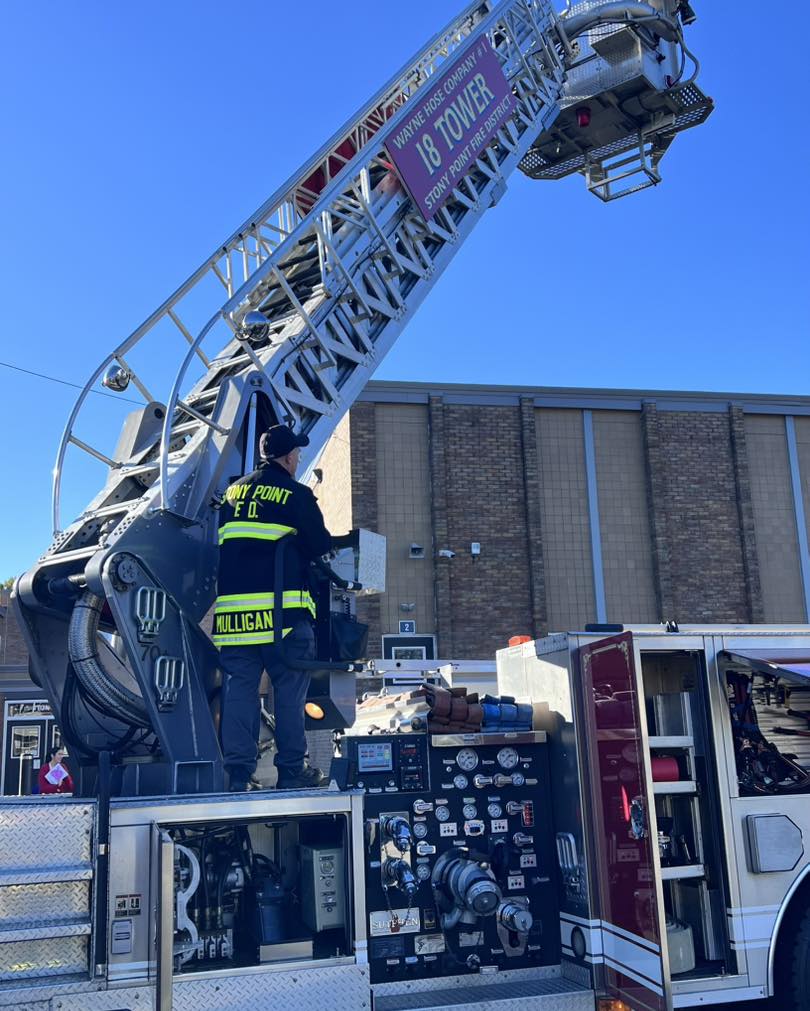 A firefighter in full gear stands on a fire truck, operating a large extended ladder labeled “Tower 18,” positioned near a brick building under a clear blue sky.