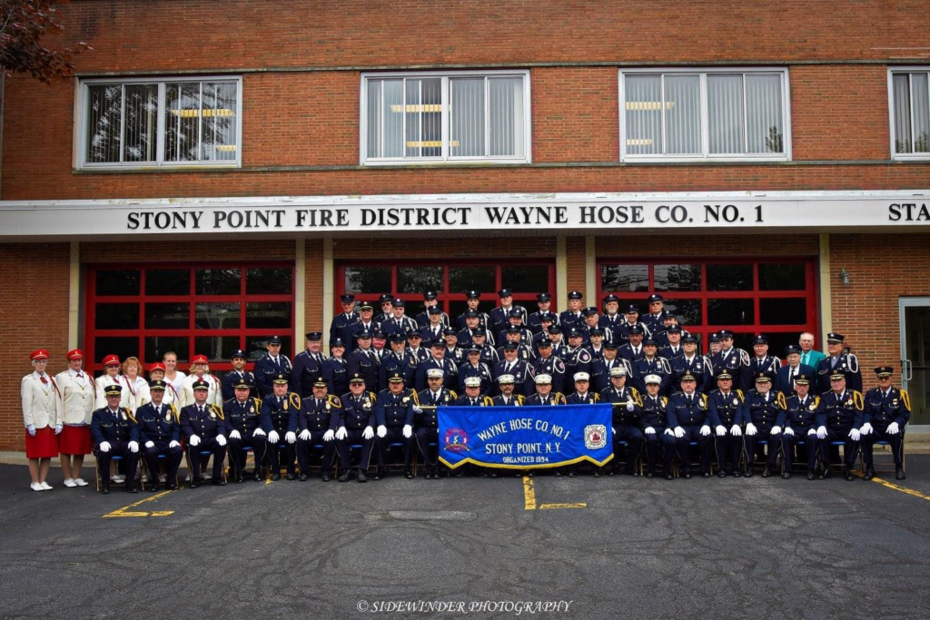 A large group of uniformed firefighters and auxiliary members pose in rows in front of the Stony Point Fire District Wayne Hose Co. No. 1 building, holding a blue company banner.