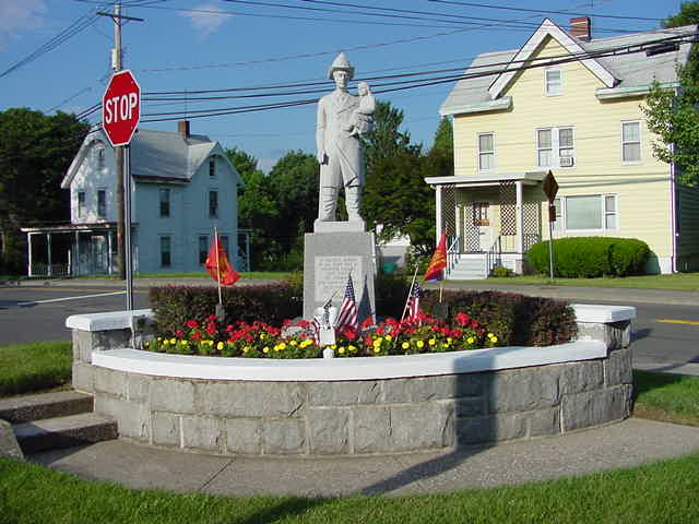 A stone firefighter statue stands on a pedestal in a small circular memorial garden with American flags, flowers, and a stop sign, surrounded by residential houses.