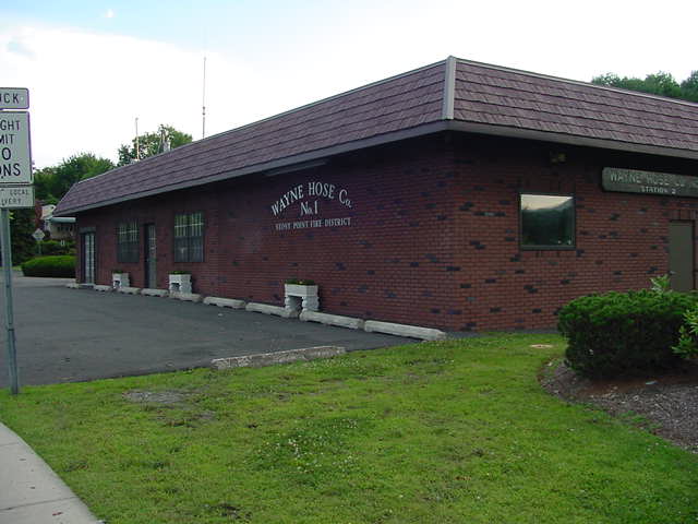 A brick building with a sign reading "Wayne Hose Co. No. 1" and "Fire District" on its side, surrounded by grass, pavement, and bushes, with a road sign partially visible in the foreground.