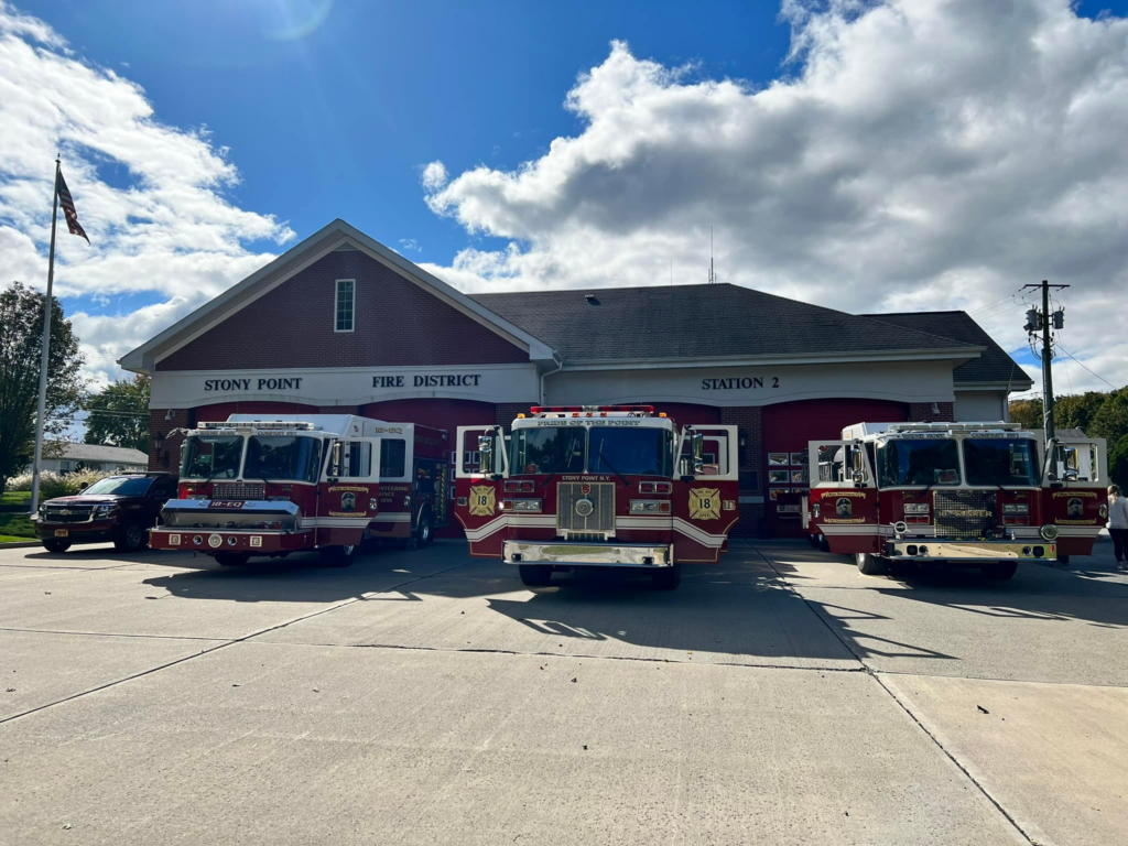 Four fire trucks are parked in front of Stony Point Fire District Station 2, a red-brick building with three garage doors. An American flag is visible to the left, under a partly cloudy sky.