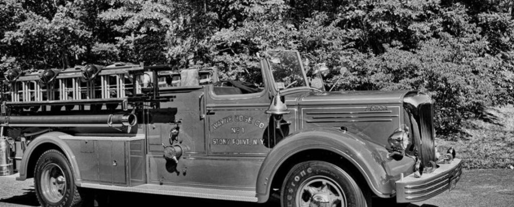 A vintage fire truck with ladders and equipment on its side is parked outdoors in front of dense trees. The vehicle has "Stony Point N.Y." and "Wayne Hose Co" written on the door.