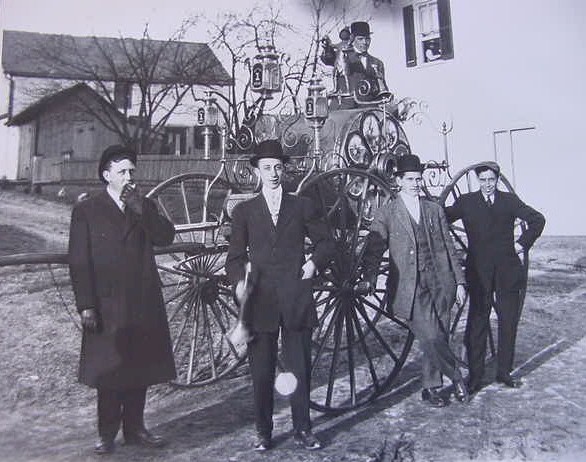 Four men in suits and hats stand in front of a vintage horse-drawn fire engine, with a man sitting on top of the vehicle; houses and bare trees are visible in the background.