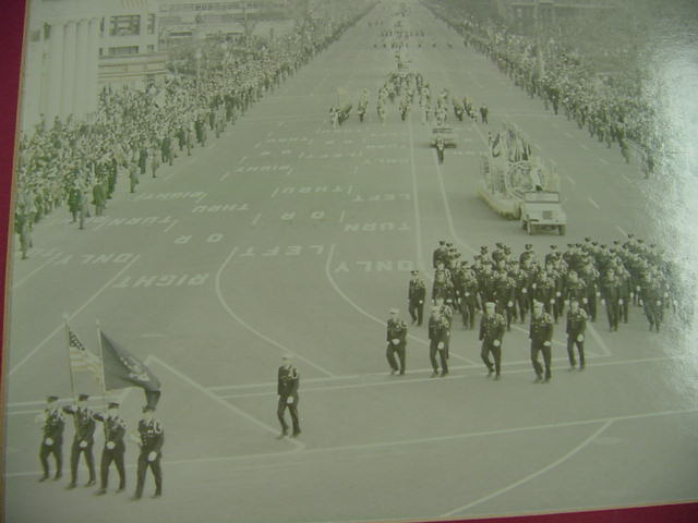 A black and white photo of a parade on a wide city street with people marching, flag bearers, a float, and crowds lining both sides of the street. Road markings indicating lanes and turns are visible.