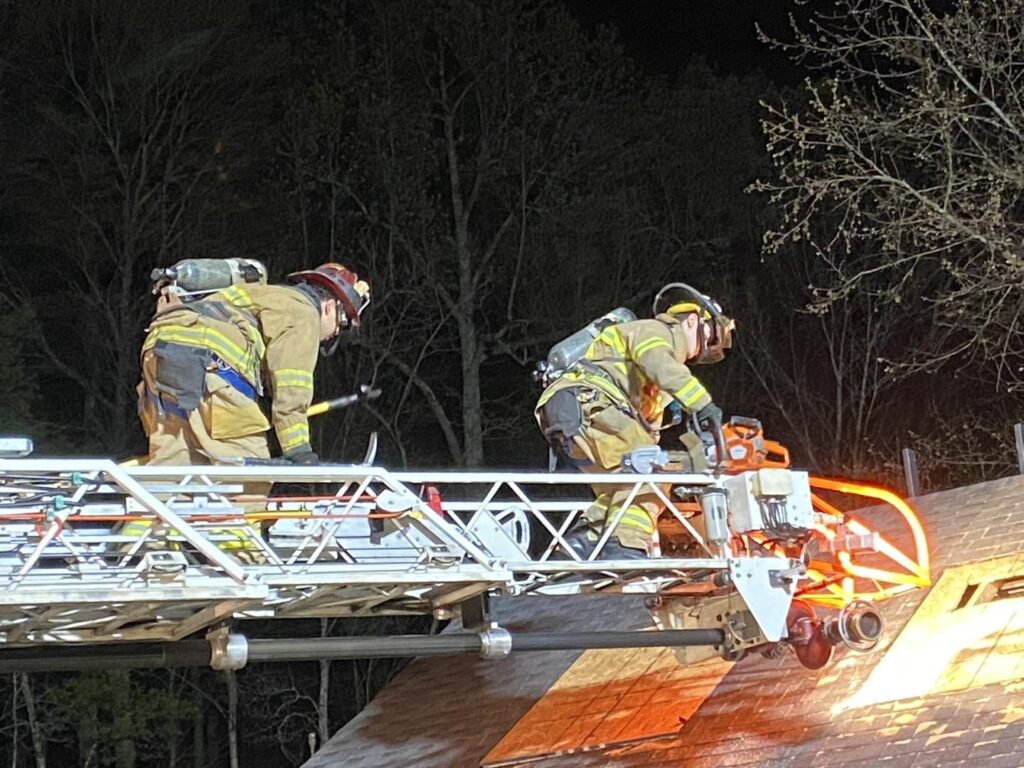Two firefighters in full gear stand on an extended ladder, using tools to cut into the roof of a building at night. Orange sparks and light are visible as one operates a power saw. Trees are visible in the background.