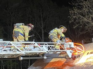 Two firefighters in full gear stand on an extended ladder, using tools to cut into the roof of a building at night. Orange sparks and light are visible as one operates a power saw. Trees are visible in the background.