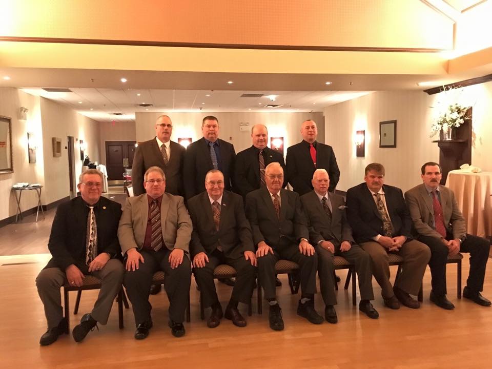 Eleven men in suits pose for a formal group photo indoors. Five are seated in front while six stand behind them. The room is warmly lit and decorated for a formal event.