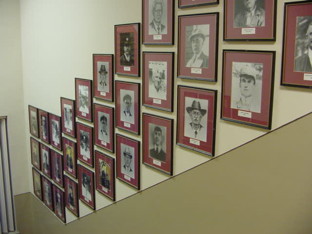 Framed black-and-white portrait photographs are displayed in rows along a stairway wall, each with a maroon mat and nameplate beneath, depicting various individuals in historical attire.